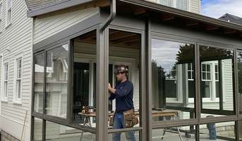 A man is focused on his work while seated at a table on a screened porch, surrounded by natural light and greenery